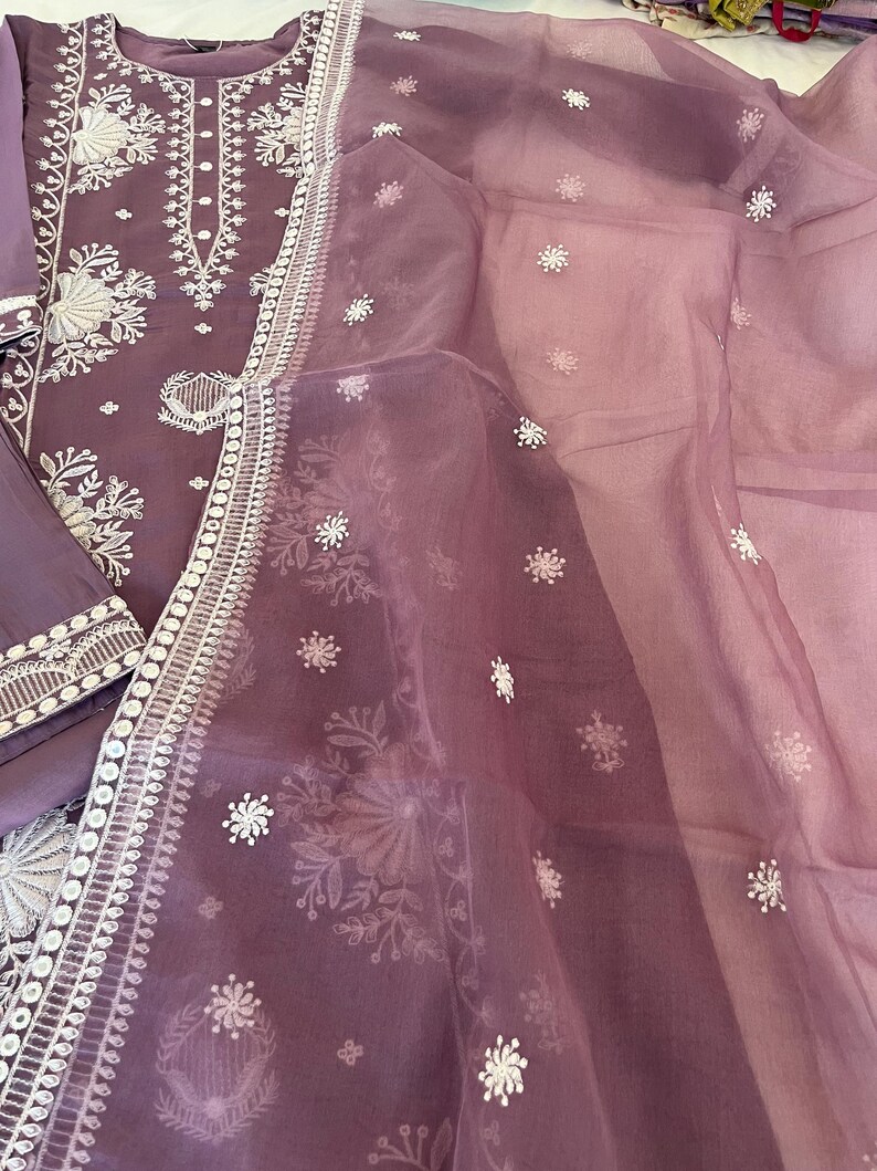 May include: A lavender-colored embroidered salwar kameez set. The tunic and pants are a solid lavender, with intricate white floral embroidery along the neckline, cuffs, and hem. A sheer, matching dupatta with scattered white floral accents completes the ensemble.