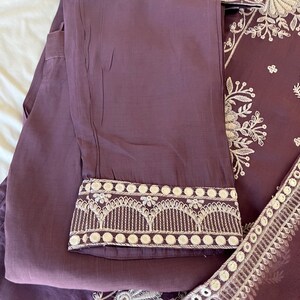 May include: A two-piece mauve outfit featuring embroidered details. The pants have a decorative hem with white embroidery. The top, partially visible, has intricate white floral embroidery. The fabric appears to be lightweight and suitable for warm weather.