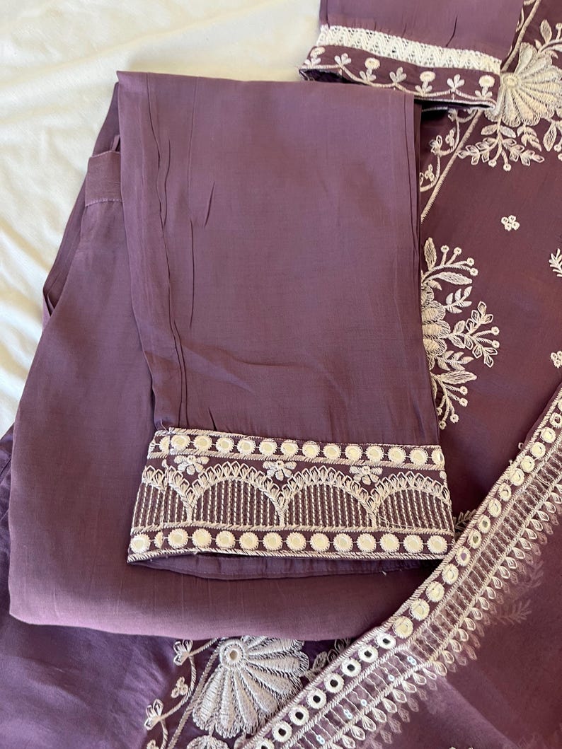 May include: A two-piece mauve outfit featuring embroidered details. The pants have a decorative hem with white embroidery. The top, partially visible, has intricate white floral embroidery. The fabric appears to be lightweight and suitable for warm weather.