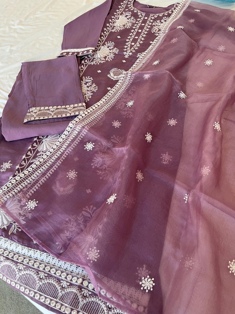 May include: A lavender-colored, embroidered three-piece outfit. The set includes a long-sleeved tunic, matching pants, and a sheer scarf. The tunic and pants feature intricate white floral embroidery along the edges and neckline.
