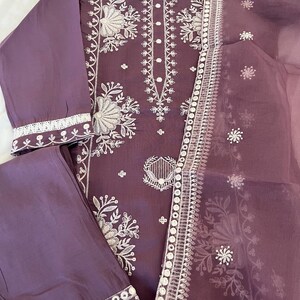 May include: A plum-colored, embroidered three-piece outfit. The set includes a long-sleeved top, matching pants, and a sheer scarf. The top and scarf feature intricate white floral embroidery along the neckline, sleeves, and edges.