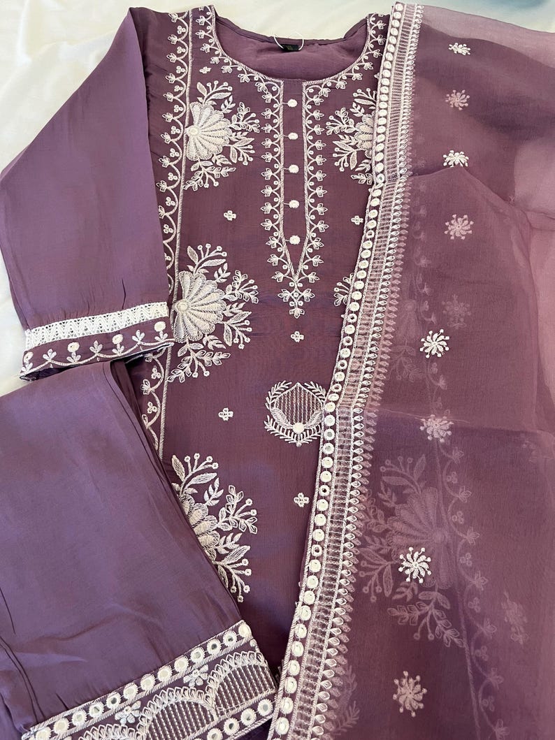 May include: A plum-colored, embroidered three-piece outfit. The set includes a long-sleeved top, matching pants, and a sheer scarf. The top and scarf feature intricate white floral embroidery along the neckline, sleeves, and edges.