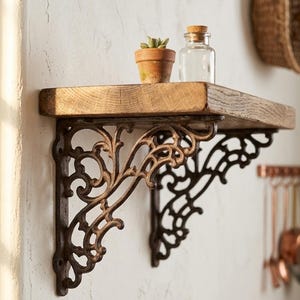 May include: A wall-mounted shelf featuring a thick, natural-toned wooden plank supported by two ornate, dark metal brackets with intricate scrollwork. A small terracotta pot containing a green succulent and a clear glass bottle sit on top.