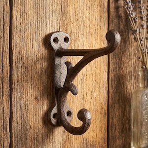 May include: A rustic, cast iron wall hook with a dark brown finish. The hook has two curved arms and a decorative flourish. It is mounted on a wooden surface, suggesting a vintage or farmhouse style.