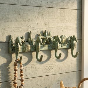 May include: A wall-mounted hook rack featuring a weathered green finish with three seahorse-shaped hooks, a central starfish, and seashell accents. The rack is designed with a horizontal bar and includes a beaded necklace hanging from the first hook.