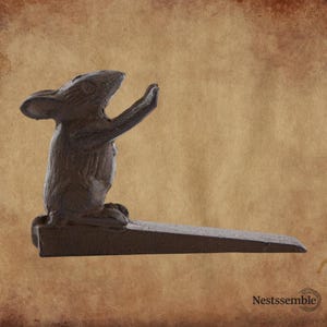 May include: A cast iron doorstop in the shape of a mouse, with its arms raised. The doorstop is dark brown and has a wedge shape for stability. The mouse is kneeling on a rectangular base.
