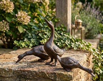 May include: Three bronze goose statues in various poses are displayed on a stone wall. One goose stands upright with its beak open, while the others are bent over. A small terracotta pot with a succulent plant sits in the foreground.