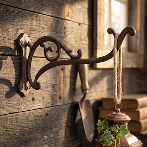 May include: A rustic, ornate, cast iron wall-mounted plant hanger. The dark brown bracket features decorative scrollwork and is attached to a weathered wooden wall. A hanging lantern with greenery is suspended from the bracket.
