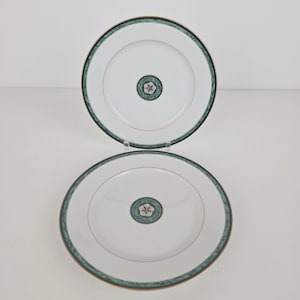 May include: Two white ceramic plates with a green and gold border. Each plate has a central design of a flower-like pattern in green, white, and gold. The plates are displayed on a white surface.
