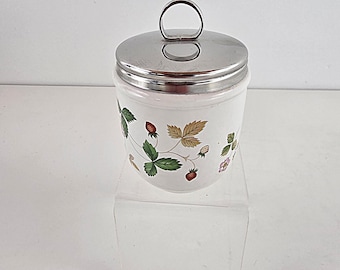 Könnte beinhalten: Ein weißer Eierbecher mit Edelstahl-Deckel und Griff. Der Becher ist mit einem Erdbeer- und Blättermuster in Rot, Grün und Hellbraun verziert. Der Eierbecher steht auf einem transparenten, schrägen Präsentationsständer.