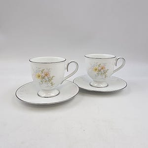 May include: Two white teacups and saucers with floral designs. Each cup has a pedestal base and a curved handle. The rims and edges of the saucers are trimmed with silver. The floral pattern includes yellow, pink, and blue flowers.