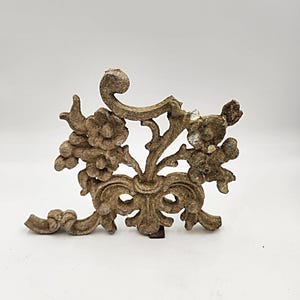 May include: A weathered, cast metal decorative architectural element featuring floral and scrollwork designs. The piece has a bow-like shape at the base and a dark, aged patina. The overall design is reminiscent of vintage or antique decor.