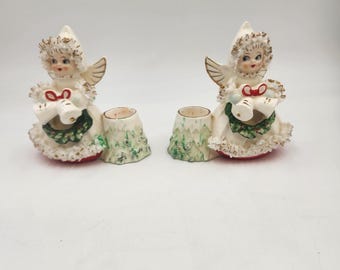 May include: A pair of vintage ceramic candle holders featuring angels holding doves and wreaths. The angels are dressed in white with gold accents and red accents on their dresses. The candle holders are in the shape of a tree stump.