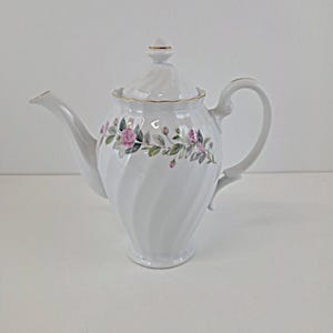 May include: White porcelain teapot with a spiral design and a lid. The teapot features a floral pattern of pink roses and green leaves. Gold trim accents the rim, handle, and spout. A classic piece for serving tea.
