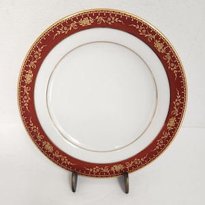 Puede incluir: Plato de cerámica blanca con borde burdeos y detalles florales dorados. El plato tiene un borde dorado y se exhibe sobre un soporte de metal. El diseño del plato presenta rosas y vides, creando una estética elegante. Este plato es adecuado para servir alimentos.