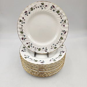 May include: A stack of ten white ceramic dinner plates with a gold rim and a floral pattern. The plates have a scalloped edge and a swirl design in the center.
