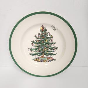 Puede incluir: Un plato de cerámica blanca con un borde verde presenta una ilustración detallada de un árbol de Navidad decorado. El árbol está adornado con adornos y coronado por Papá Noel. Los regalos están en la base del árbol. Un copo de nieve está a la derecha.