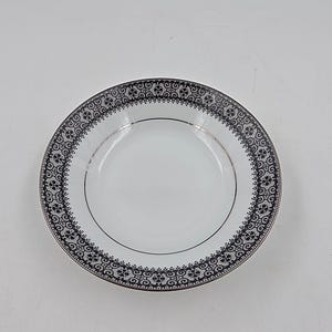 May include: White ceramic bowl with a black and silver floral pattern around the rim. The bowl has a gold-colored trim and a simple, elegant design. A classic piece for serving soups, salads, or desserts.