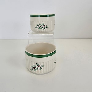 Puede incluir: Dos ramequines de cerámica blanquecina con una banda verde alrededor del borde y un diseño de acebo. Los ramequines tienen detalles verticales acanalados y están apilados sobre un soporte acrílico transparente. El diseño de acebo presenta hojas verdes y bayas rojas.