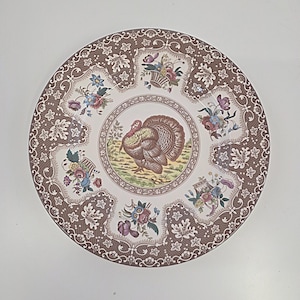 May include: Round ceramic plate with a turkey in the center, surrounded by floral and basket designs. The rim features a brown and white pattern. The plate has a cream-colored background and is suitable for serving food.