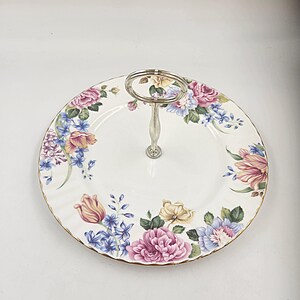 Peut inclure: Un support à gâteau en céramique blanche avec une poignée argentée. Le support est décoré d'un motif floral dans des tons de rose, jaune, bleu et vert.