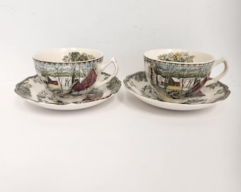 Può includere: Due tazze da tè e piattini vintage con un design a tema invernale. Le tazze e i piattini sono bianchi con un motivo marrone, verde e rosso. La scena rappresenta un paesaggio innevato con case e alberi.