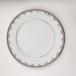Könnte beinhalten: Ein weißer Keramik-Dinnerteller mit silbernem Rand und einem zarten Blumenmuster in Rosa- und Grüntönen.