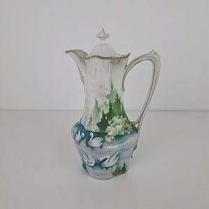 Pode incluir: Uma cafeteira de porcelana branca e dourada com tampa. Apresenta uma paisagem pintada com árvores verdes, cisnes brancos e um lago azul. A alça e o bico são detalhados com acabamentos dourados.