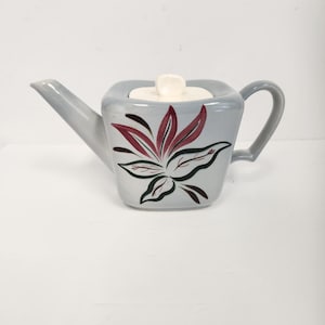 May include: A grey ceramic teapot with a square body and a white lid. The teapot features a floral design in shades of red, green, and black.