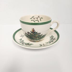 Puede incluir: Un juego de taza y platillo blancos con borde verde. La taza presenta un diseño de árbol de Navidad con una figura en la parte superior. El platillo tiene un borde verde a juego y detalles de hojas de acebo. Ideal para celebraciones navideñas.