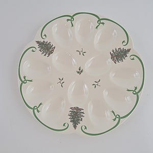 Peut inclure: Un plat de service à œufs rond en céramique blanc cassé comportant douze alvéoles ovales. L'assiette est ornée d'une bordure en ruban vert, de petits brins de houx et de deux illustrations détaillées de conifères avec des pommes de pin.