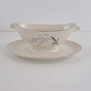 May include: A cream-coloured ceramic gravy boat with an attached oval underplate, featuring a delicate grey botanical illustration of wheat stalks on the side and a thin silver metallic trim along the edges of both the boat and the plate.
