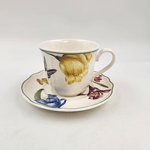 Puede incluir: Un juego de taza y platillo de té de porcelana blanca con un diseño floral con flores amarillas, azules y moradas y una mariposa. La taza y el platillo tienen un borde verde.