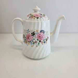 May include: White ceramic teapot with a floral design featuring pink roses and other flowers. The teapot has a lid with a small finial and a curved handle. The body has a spiral design.