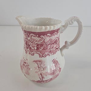 Peut inclure: Un pichet à crème en céramique avec une base blanche et un motif de transfert rose foncé. Le design comprend une bordure florale sur le haut et une scène paysagère avec des bâtiments et des arbres sur le corps. Il possède un bord festonné.