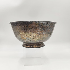 May include: A silver-toned trophy bowl with a dark, aged patina. The bowl has a wide, rounded shape and a small, circular base. Engraved text is visible around the bowl's rim, indicating an award. The trophy sits on a clear, rectangular stand.