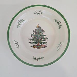 May include: White ceramic serving bowl with a green rim and a Christmas tree design in the center. The tree is decorated with ornaments and surrounded by wrapped gifts. Sprigs of holly and mistletoe are also featured.