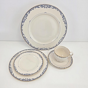 May include: A cream-coloured dinnerware set with blue and gold detailing. The set includes a large dinner plate, a smaller plate, a saucer, and a teacup. The plates and saucer have a blue floral border and a gold rim. The teacup has a gold rim.