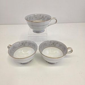 Könnte beinhalten: Drei Porzellan-Teetassen mit hellgrauer Außenseite und weißer Innenseite. Jede Tasse hat einen goldenen Rand und Henkel, mit einem Blumendesign am Rand. Eine Tasse steht auf einem transparenten Ständer.