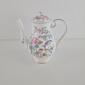 May include: A white porcelain teapot featuring a colorful floral and bird pattern with pink, red, blue, and green details. The teapot has a curved handle, a long spout, a matching lid with a small finial, and gold trim along the edges.