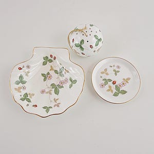 May include: A trio of white ceramic items featuring a strawberry pattern. The set comprises a shell-shaped dish, a small round plate, and a spherical tea infuser. Each piece has a gold edge and is adorned with green leaves, pink flowers, and red strawberries.