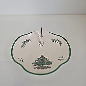 May include: A white ceramic serving dish with a green trim and a unique shape. The dish features a Christmas tree design in the center, with holly and berry accents. The dish measures approximately 10 inches in diameter.