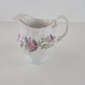 Può includere: Una lattiera in porcellana bianca con bordo smerlato e finiture color oro. Il corpo è decorato con un motivo floreale di rose rosa e foglie verdi. Presenta un manico curvo e un beccuccio per versare, poggiante su una base a piedistallo.
