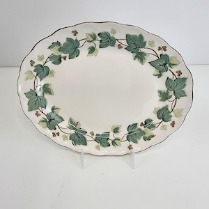 Puede incluir: Una fuente ovalada con borde festoneado. La fuente de cerámica blanca presenta un diseño de hojas de hiedra verde alrededor del borde y un borde marrón. La fuente está sostenida por soportes de plástico transparente.