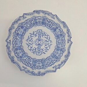 May include: A white ceramic plate with a blue floral design. The plate has a scalloped edge and a detailed pattern of flowers and leaves. The design is symmetrical and covers the entire surface of the plate.