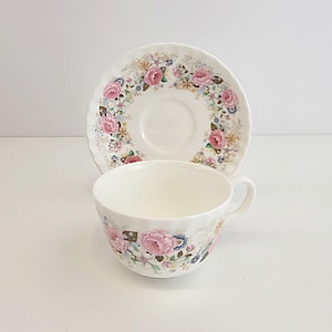 May include: A white teacup and saucer set. The cup and saucer are decorated with a floral pattern featuring pink roses, blue and yellow flowers, and green leaves. The set is on a white background.
