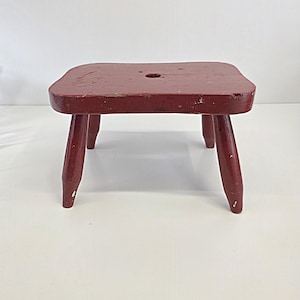 May include: A small, square, red wooden stool with four angled legs. The stool has a slightly distressed finish, with visible wear and tear on the surface. A small hole is centered on the top.