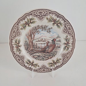 Puede incluir: Plato decorativo con una escena de faisanes. El plato presenta un borde marrón y blanco con diseños florales y de bellotas. El centro representa dos faisanes en un paisaje con un árbol, una valla y colinas.