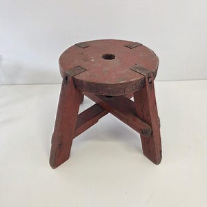 May include: A small, round, red wooden stool with a circular seat and four angled legs. The seat has a hole in the center and is reinforced with metal brackets. The stool has a rustic, vintage appearance.