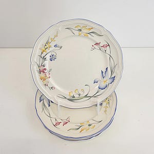 May include: A stack of white ceramic plates with a scalloped edge and a blue rim. Each plate is decorated with a floral design featuring yellow, pink, and blue flowers. The plates are stacked on a clear plastic stand.